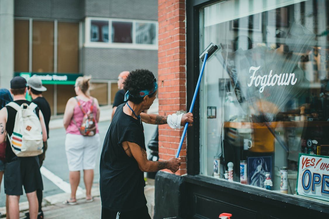 The image shows a person cleaning the window of a store while a group of people stands nearby.